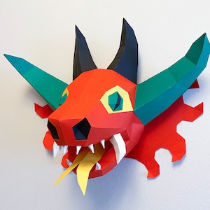 May include: A red, green, and black paper dragon head sculpture with a yellow tongue sticking out. The dragon has a large, toothy mouth and a single green eye.