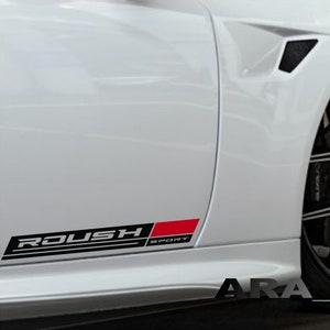 Puede incluir: Un coche blanco con una calcomanía "ROUSH SPORT" negra y roja en el lateral. El coche tiene ruedas negras con un diseño de radios plateados.