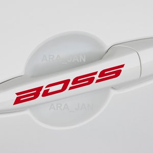 Può includere: Maniglia della portiera dell'auto bianca con un adesivo rosso "BOSS". La maniglia ha un piccolo buco della serratura e un rivestimento in plastica bianca con il testo "ARA_JAN" su di esso.