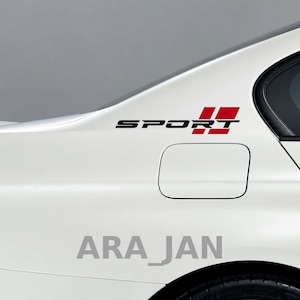 Puede incluir: Un coche blanco presenta una pegatina negra y roja en el panel trasero. La pegatina muestra la palabra SPORT en letras mayúsculas negras, seguida de dos bloques rectangulares rojos. El diseño está situado sobre la tapa del depósito.