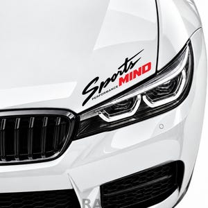 May include: A white car hood features a black and red vinyl decal sticker with the text Sports PERFORMANCE MIND. The decal is positioned diagonally above the passenger side headlight, which has a distinct angular LED light signature.