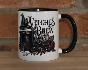 May include: White ceramic mug with a black rim and handle. The mug features a black and green illustration of a witch with a cauldron and the text "Witches Brew Drink Co. Quality Potions Since 1913."