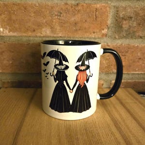 Könnte beinhalten: Weiße Keramiktasse mit schwarzem Rand und Henkel. Die Tasse zeigt ein Design mit zwei Hexen in schwarzen Kleidern und Hüten, die Regenschirme halten, um die herum Fledermäuse fliegen.