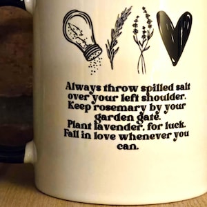 May include: White ceramic mug with a black rim and handle. The mug has a black and white illustration of a salt shaker, rosemary, lavender, and a heart. The text reads: "Always throw spilled salt over your left shoulder. Keep rosemary by your garden gate. Plant lavender, for luck. Fall in love whenever you can."