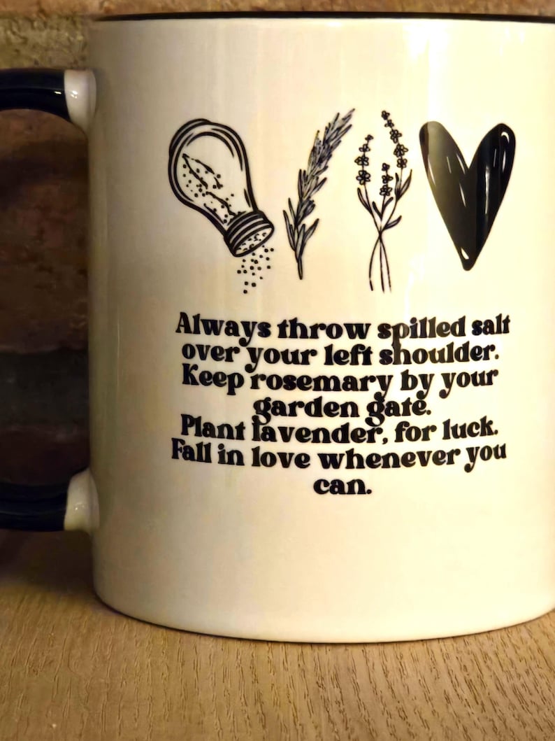 May include: White ceramic mug with a black rim and handle. The mug has a black and white illustration of a salt shaker, rosemary, lavender, and a heart. The text reads: "Always throw spilled salt over your left shoulder. Keep rosemary by your garden gate. Plant lavender, for luck. Fall in love whenever you can."