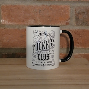 May include: White ceramic coffee mug with a black rim and handle. The mug has a black graphic that says "Grumpy Fuckers Club - 100% Bitter, Jaded & Proud - Lifetime Member".