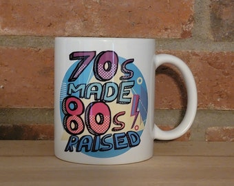 May include: White ceramic mug with a colorful graphic design that says "70s Made 80s Raised".