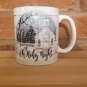 May include: White ceramic mug with a black and white image of a church and trees. The mug has the text "Oh Holy Night" written on the bottom.