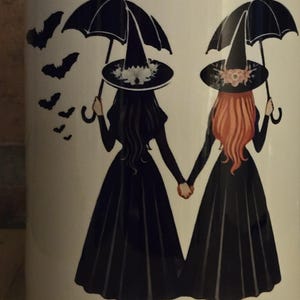 May include: White ceramic mug with a black rim. The mug features a graphic of two witches holding hands, each with a black umbrella and wearing a black witch hat. One witch has black hair and the other has red hair. There are several black bats flying around the witches.