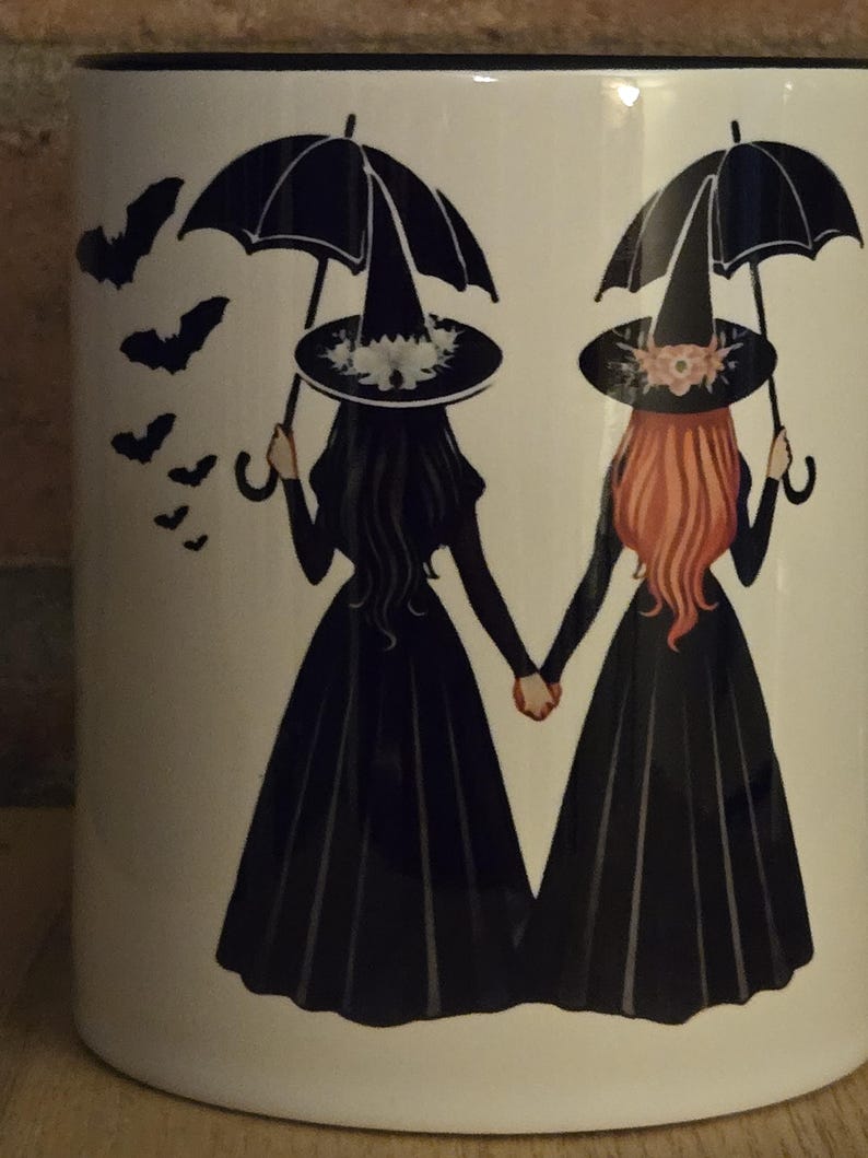 May include: White ceramic mug with a black rim. The mug features a graphic of two witches holding hands, each with a black umbrella and wearing a black witch hat. One witch has black hair and the other has red hair. There are several black bats flying around the witches.