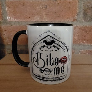 May include: White ceramic mug with a black rim and handle. The mug features a black and white graphic of a gothic archway with a bat, the words "Bite Me" and a red dripping vampire mouth.