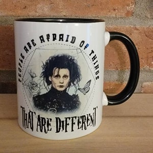 May include: White ceramic mug with a black rim and handle. The mug features a black and white illustration of a character from the movie Edward Scissorhands. The text on the mug reads "People are afraid of things that are different."