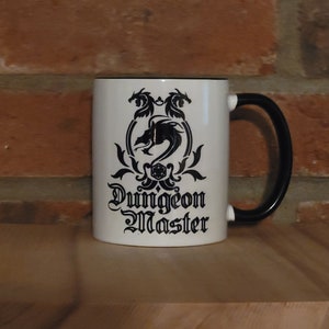 May include: White ceramic mug with a black rim and handle. The mug has a black and white design of a dragon with the words "Dungeon Master" printed below.