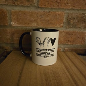 May include: White ceramic mug with a black handle and black rim. The mug has a black design with a salt shaker, rosemary sprigs, and a heart. The text on the mug reads: "Always throw spilled salt over your left shoulder. Keep rosemary by your garden gate. Plant lavender, for luck. Fall in love whenever you can."