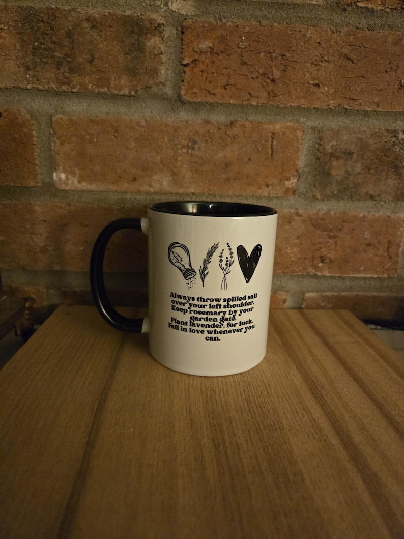 May include: White ceramic mug with a black handle and black rim. The mug has a black design with a salt shaker, rosemary sprigs, and a heart. The text on the mug reads: "Always throw spilled salt over your left shoulder. Keep rosemary by your garden gate. Plant lavender, for luck. Fall in love whenever you can."