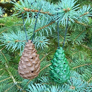 May include: Two pine cone ornaments, one brown and one green, hang from black cords against a backdrop of evergreen branches. The ornaments have a textured surface and are designed with holes.