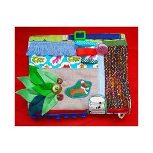 Pode incluir: Um livro de tecido multitexturizado e multicolorido com várias texturas e objetos para os bebês e crianças pequenas explorarem. O livro apresenta uma folha de feltro verde, um dachshund de feltro marrom, uma franja de feltro azul, uma fita vermelha, uma fivela de plástico verde e um botão azul.