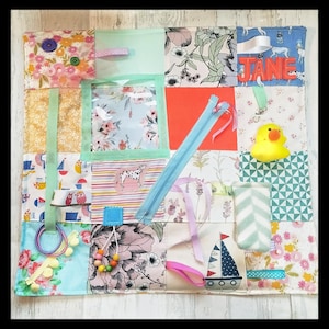 Op de afbeelding: Een kleurrijk sensorisch activiteitenbord met diverse texturen en interactieve elementen. Voorzien van een rits, linten, knopen, een badeend en een naamlabel met "JANE". Het bord is gemaakt van stoffen vierkanten met bloemen-, dieren- en nautische patronen.