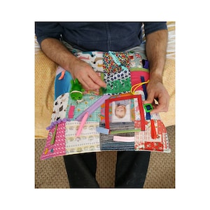 以下が含まれることがあります： さまざまなテクスチャとインタラクティブな要素を備えた、カラフルな感覚アクティビティマット。写真、ジッパー、リボン、緑のカエルのおもちゃ、バックルが特徴です。マットはさまざまなパターンと色のパッチワーク生地で作られています。