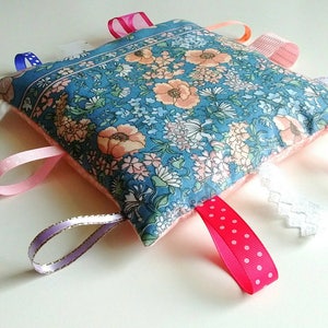 May include: A blue and pink fabric square with various textures and colors attached. The square has a floral pattern and is filled with soft stuffing. The textures include a white rope, pink ribbon, purple ribbon with gold trim, white lace, and a red ribbon with white polka dots.