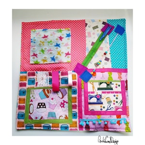 May include: A pink and blue fabric activity mat with various textures and features for babies and toddlers. It includes a zipper, buttons, ribbons, a pocket, and a variety of fabrics with different patterns. The mat is labeled "CushiCushi Designs".