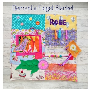 Puede incluir: Una manta sensorial para la demencia con varios elementos interactivos. Incluye botones, cintas, una flor, una aplicación de pájaro y el nombre "ROSE". La manta está hecha de telas de patchwork coloridas.