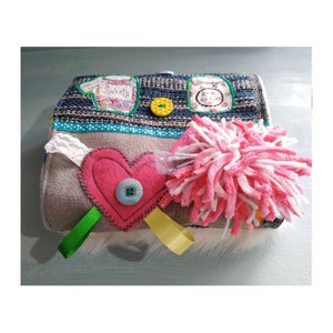 Op de afbeelding: Een grijze en blauwe stoffen tas met een roze hart, een roze en witte pompon en een groen en geel lint. De tas heeft verschillende texturen en patronen, waaronder een gestreepte stof, een kantversiering en een knoop.