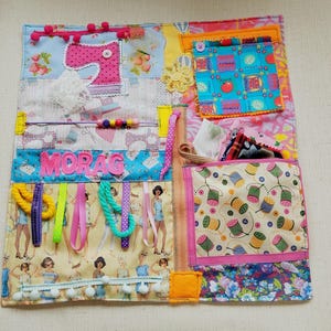 May include: A colorful, handmade sensory activity board with various textures and interactive elements. Features fabric patches with sewing-themed patterns, ribbons, buttons, and the name "MORAG". Includes a pocket and decorative trim.