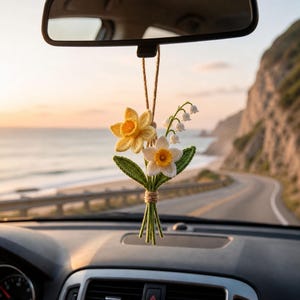 May include: A handmade crochet car charm featuring a bouquet of yellow daffodils, white daffodils, and lily of the valley flowers. The flowers are tied together with twine and hang from a rearview mirror. The background shows a coastal road and ocean.