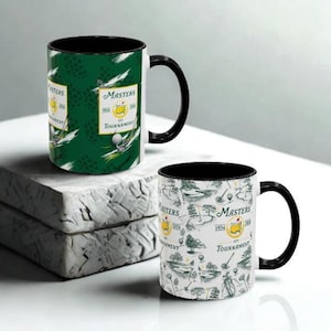 May include: Two ceramic coffee mugs with black handles and interiors. One mug features a green background with golf-themed graphics and the text "Masters Tournament 1934". The other mug has a white background with golf course illustrations and the same text.