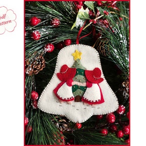 Op de afbeelding: Een klokvormig viltornament met twee figuren in rode mutsen en witte schorten voor een groene kerstboom met een gele ster. Het ornament heeft een crèmekleurige achtergrond met zichtbare stiksels langs de randen en een rood lint.