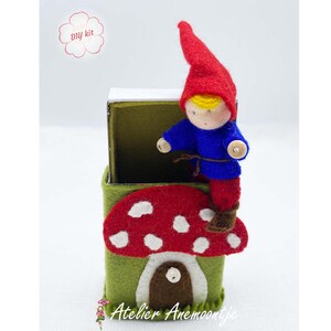 May include: DIY kit for a felt gnome doll house. The house is a green matchbox with a red and white mushroom roof. The gnome doll is wearing a blue shirt, red pants, and a red hat. The text "DIY kit" and "Atelier Anemoonkje" are visible on the image.