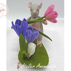 May include: A gray felt mouse holding a pink felt flower, standing in front of a felt flower arrangement with purple and white flowers. The flowers are on a green felt base. The image includes the text "DIY kit".