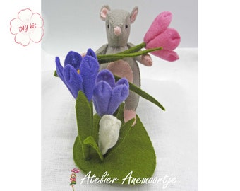 May include: A gray felt mouse holding a pink felt flower, standing in front of a felt flower arrangement with purple and white flowers. The flowers are on a green felt base. The image includes the text "DIY kit".