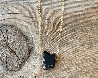 May include: A gold chain necklace with a black obsidian stone pendant. The stone is rough and has a natural shape. The necklace is on a wooden background with the text "Black Obsidian Protection Stone".
