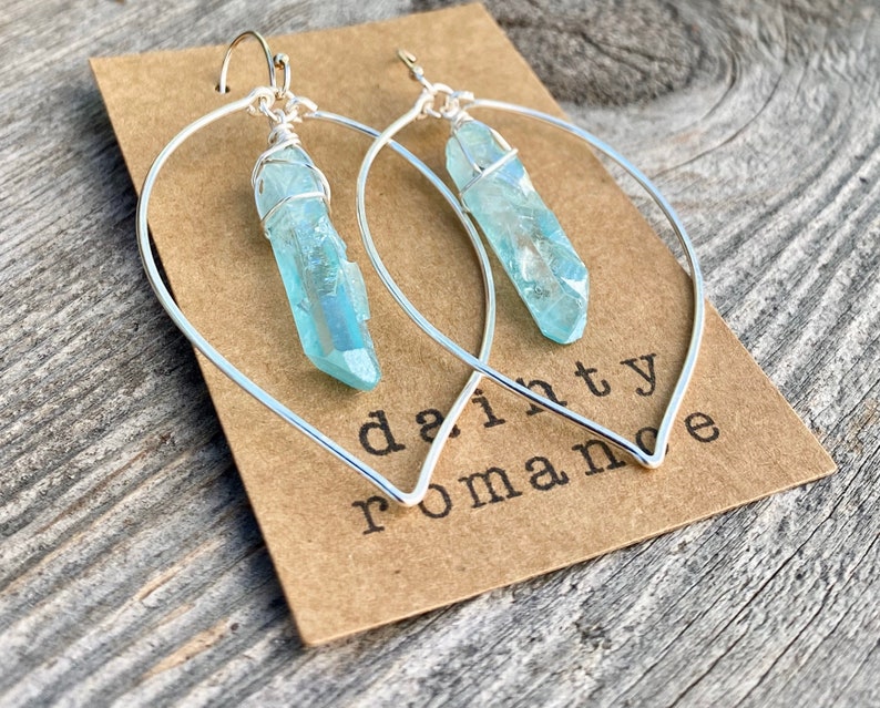 Puede incluir: Aretes de aro de plata con un colgante de cristal azul claro. Los aretes est&aacute;n en una tarjeta marr&oacute;n con el texto "dainty romance".