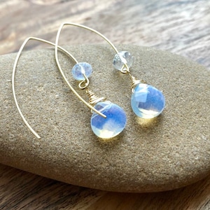 May include: A pair of gold wire earrings with teardrop-shaped opal stones. The stones are a pale blue color with a slight iridescent shimmer.
