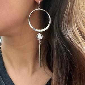 May include: Silver hoop earrings with a starburst charm and a long, thin bar hanging from it.