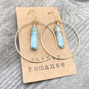 Puede incluir: Aretes de aro dorados con un colgante de cristal azul claro que cuelga de cada aro. Los aretes est&aacute;n sobre un fondo marr&oacute;n con el texto "daiby romance" visible.