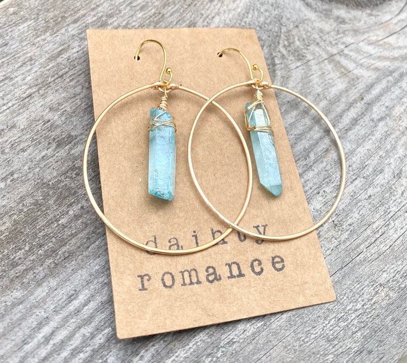 Puede incluir: Aretes de aro dorados con un colgante de cristal azul claro que cuelga de cada aro. Los aretes est&aacute;n sobre un fondo marr&oacute;n con el texto "daiby romance" visible.