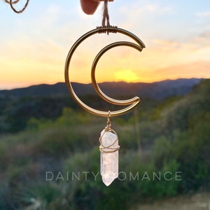 May include: Gold crescent moon pendant with a clear quartz crystal hanging from it. The pendant is on a string and being held by a hand. The background is a sunset over a mountain range.
