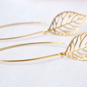 May include: A pair of gold-colored leaf-shaped earrings. Each earring features a delicate, openwork leaf design suspended from a curved wire. The earrings are presented against a soft, neutral background, highlighting their intricate details.