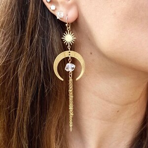 May include: Gold crescent moon earrings with a star charm and a crystal dangle. The earrings have a delicate chain that hangs below the crystal.