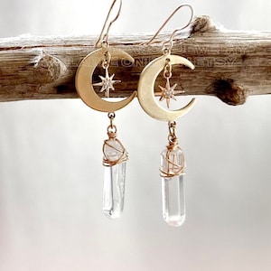 May include: Gold crescent moon earrings with a small star charm and a clear quartz crystal pendant hanging from each moon. The crystals are wrapped in gold wire.