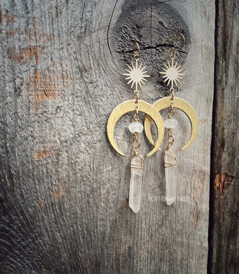 Pu&ograve; includere: Orecchini a forma di mezzaluna dorati con un ciondolo a stella e un pendente di cristallo di quarzo trasparente. Gli orecchini sono su uno sfondo di legno.