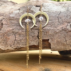 May include: Gold crescent moon earrings with a clear crystal charm and multiple gold chains dangling from the bottom.