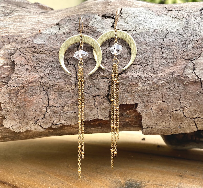 May include: Gold crescent moon earrings with a clear crystal charm and multiple gold chains dangling from the bottom.