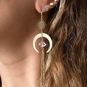 May include: Gold crescent moon earrings with a dangling crystal and a star charm. The earrings are hanging from a person's ear.