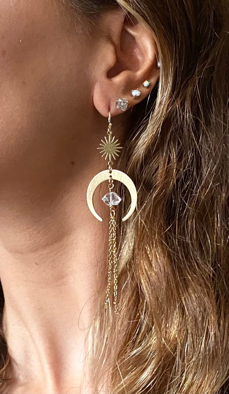 May include: Gold crescent moon earrings with a dangling crystal and a star charm. The earrings are hanging from a person's ear.