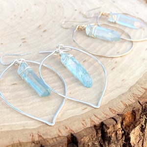 Puede incluir: Un par de pendientes de aro de plata con un colgante de cristal azul claro que cuelga de cada aro. Los aros son ligeramente ovalados y tienen una parte inferior puntiaguda.